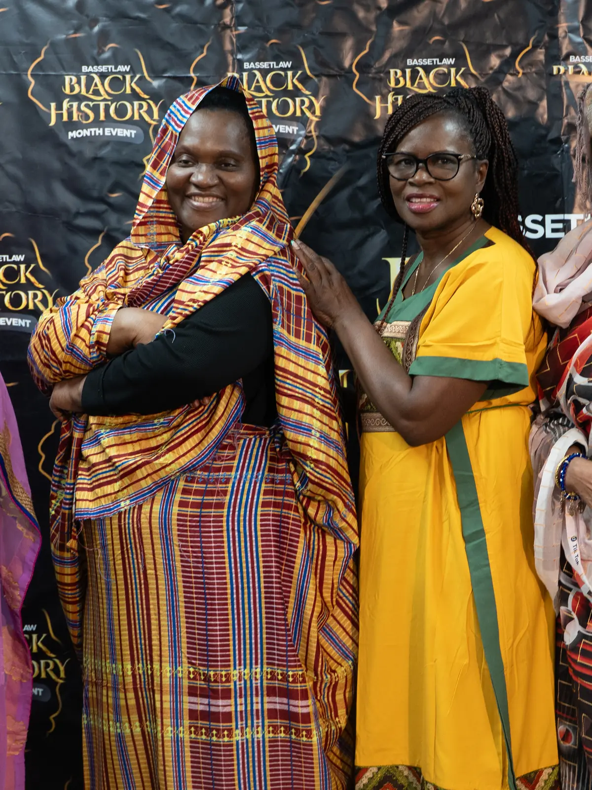 Two women in colourful outfits, one in a yellow dress, smiling at a Black History Month gathering.